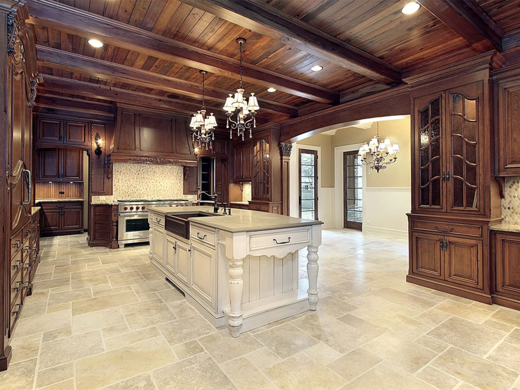Spacious kitchen with rustic charm featuring rich dark wood cabinetry, a large central island with a light-colored countertop, terracotta tile flooring, and exposed wooden beams on the ceiling. Modern appliances are nestled within the cabinets, and elegant chandeliers provide lighting. An open doorway leads to an adjoining room with light walls and additional lighting fixtures